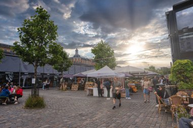 Kopenhag 'da canlı bir pazar, beyaz ve gri gölgelerin altında tezgahlar, alışveriş ve yemek yapan insanlar, ve günbatımında arka planda yükselen bir bina..