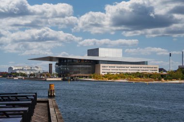 Kopenhag Opera Binası, deniz kenarında yer alır, üzerinde kantilli bir çatı, ön planda ahşap bir iskele ve uzak kentsel yapılar bulunur..