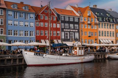 Danimarka 'nın Kopenhag kentindeki Nyhavn bölgesinde canlı tarihi binalar, SA 98 damgalı beyaz bir tekne ve pembe bir akşam gökyüzü altında açık kafeler bulunmaktadır..