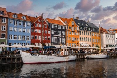 Danimarka 'nın Kopenhag kentindeki Nyhavn Kanalı' nda canlı yüzler, rıhtımlı tekneler, açık hava koltukları ve bulutlu bir gökyüzünün altında hareketli bir gezinti alanı vardır..