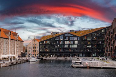 Kopenhag rıhtımında siyah eğimli bir çatı, kırmızı fayanslı tarihi yapılar, küçük tekneler ve canlı kırmızı ve turuncu bir gökyüzü bulunur..