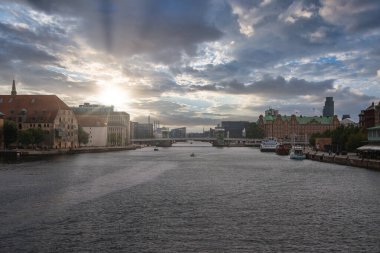 Kopenhag, Danimarka 'da bir kanal, gün batımında kırmızı çatılı ve yeşil çatılı binalar, uzak bir köprü, küçük tekneler ve gökyüzünde dramatik güneş ışınları..