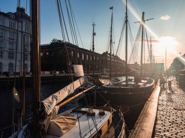 Kopenhag, Danimarka 'da bir kanal, gün batımında demirli yelkenliler, beyaz Avrupa tarzı bir bina ve arka planda kemerli tuğla bir yapı..