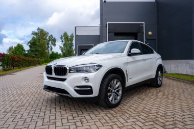 Büyük bir garaj kapısı, yeşillik ve bulutlu gökyüzü olan koyu gri endüstriyel bir binanın yanındaki asfalt bir yolda beyaz bir BMW X6 'ya yakın dur..