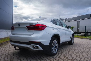Asfaltlı bir yüzeye park edilmiş beyaz bir BMW X6, eğimli çatı çizgisi, çift egzozu ve X6 rozeti sergiliyor. Endüstriyel binalar ve bulutlu bir gökyüzü görülebilir.