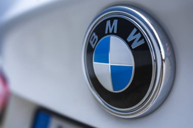 Mavi ve beyaz çeyrekli BMW logosu, bulanık bir arka plan ve plakanın bir parçası olan beyaz bir BMW X6 'nın arkasına keskin bir şekilde odaklanmıştır..