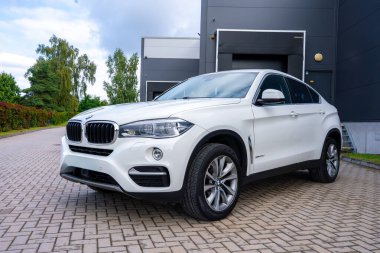 Pürüzsüz tasarım detayları olan beyaz bir BMW X6 asfalt yola park edilmiş. Sahnede modern gri bir bina, çitler, ağaçlar ve bulutlu bir gökyüzü yer alıyor..