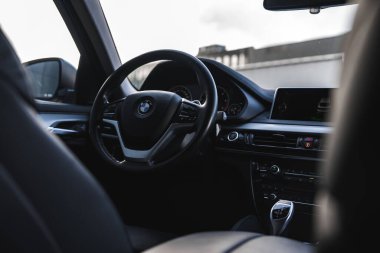 Bir BMW X6 'nın iç görüntüsü direksiyona, gösterge paneline ve dişli seçiciye odaklanıyor. Bulanık kentsel bir ortam ve bir bina dışarıda görülebilir..