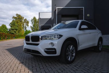 Ağaçlar ve çitlerle çevrili büyük bir garaj kapısı olan koyu gri endüstriyel bir binanın önündeki asfalt bir yolda beyaz bir BMW X6 park edilmiştir..