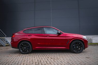 Parlak kaplamalı kırmızı bir BMW X4 ve asfalt bir yüzeye park edilmiş siyah alaşımlı tekerlekler. Arka planda metal merdivenleri olan modern gri endüstriyel bir bina var..