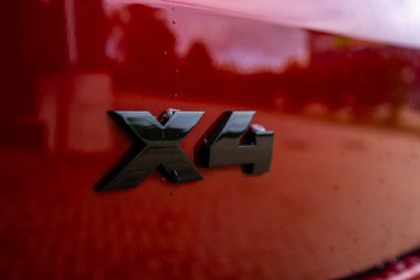 Kırmızı bir BMW X4 'ün arkasındaki X4 rozetinin yakın görüntüsü. Parlak siyah amblem kırmızı boyayla çelişiyor, ağaçları ve su damlacıklarıyla yapıları yansıtıyor..