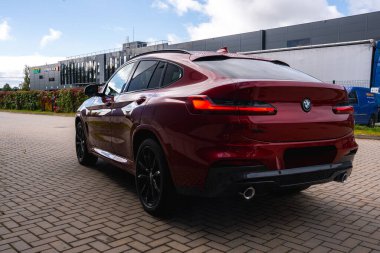 Kırmızı bir BMW X4 asfalt bir yüzeye park edilmiş, eğimli çatı çizgisi ve çift egzozları sergileniyor. Arka planda modern bir bina ve yeşillik görülebilir..