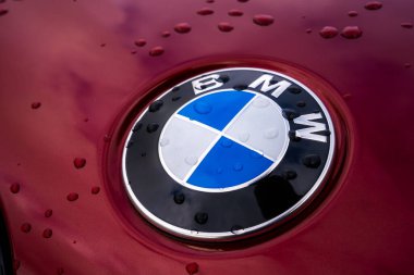 Kırmızı bir BMW aracının kaputundaki BMW logosunun yakın görüntüsünde su damlacıkları ve parlak yüzeyde soluk gökyüzü yansımaları var..
