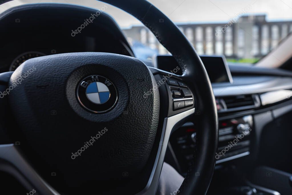 Interior view of a BMW X6 focusing on the steering wheel with BMW logo, dashboard, and infotainment screen. A multi story building is visible outside.