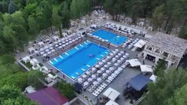 Birçok şemsiyesi, güneşliği ve yaz gününün tadını çıkaran insanlarla dolu büyük bir açık havuz kompleksi olan hava aracı, yeşil ormanlarla çevrili modern tatil beldesini gösteriyor..