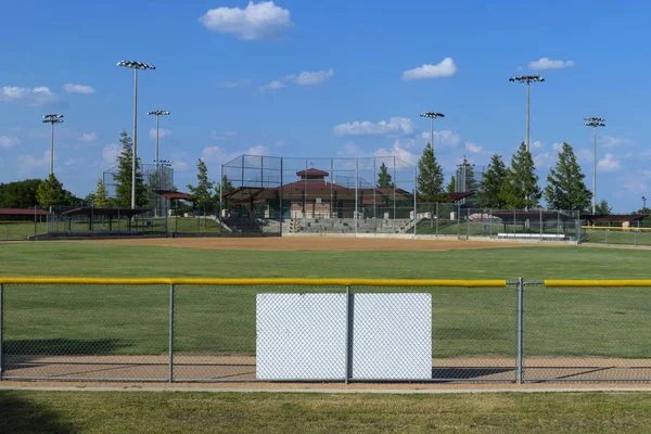 View of an empty baseball field from a hill overlooking the fence in centerfield with the diamond, bases, dugouts, and activity buildings in the distance, while fluffy, white clouds drift by in the bright, blue sky. The blank, white back of a sign is