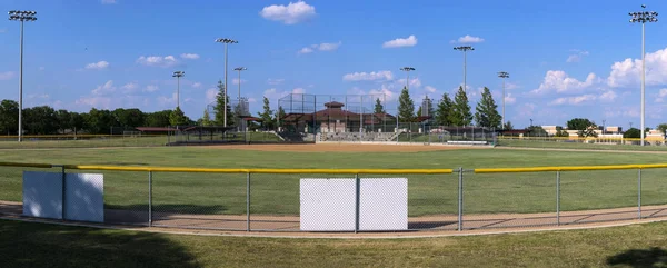 Panoramic view of an empty baseball field from a hill overlooking the fence in centerfield with the diamond, bases, dugouts, and activity buildings in the distance, while fluffy, white clouds drift by in the bright, blue sky. The blank, white back of