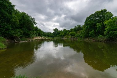 Sakin bir nehrin kıyısında uzanan ağaçlar, sakin bir yaz sabahı gökyüzünü dolduran bulutlarla birlikte suya yansıyor..