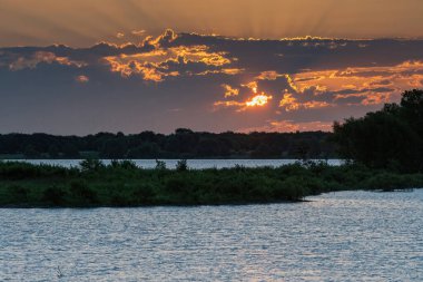 Güneş, sabahın erken saatlerinde ağaçlarla kaplı bir yarımadanın üzerindeki bulutların arasından süzülerek suları iki göle ayırıyor..
