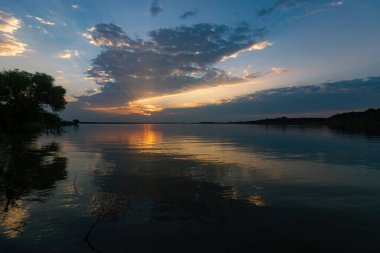 Güneşin sabahın erken saatlerinde doğduğu sahilde bulutları, güneş ışığını ve ağaçları yansıtan sakin, cam gibi bir gölün suyu..