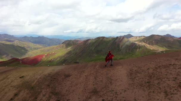Aerial drone disparo de una chica en un auténtico poncho de arco iris senderismo en una colina de montaña. Valle Rojo de los Andes, Montaña de colores, Perú — Vídeos de Stock