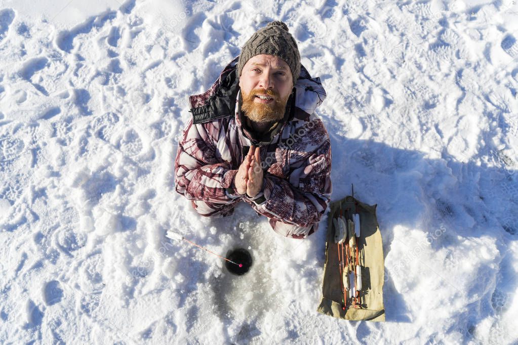 Bearded european sad man is praying and appealing to the god while he getting fail at the winter fishing from ice hole on the river