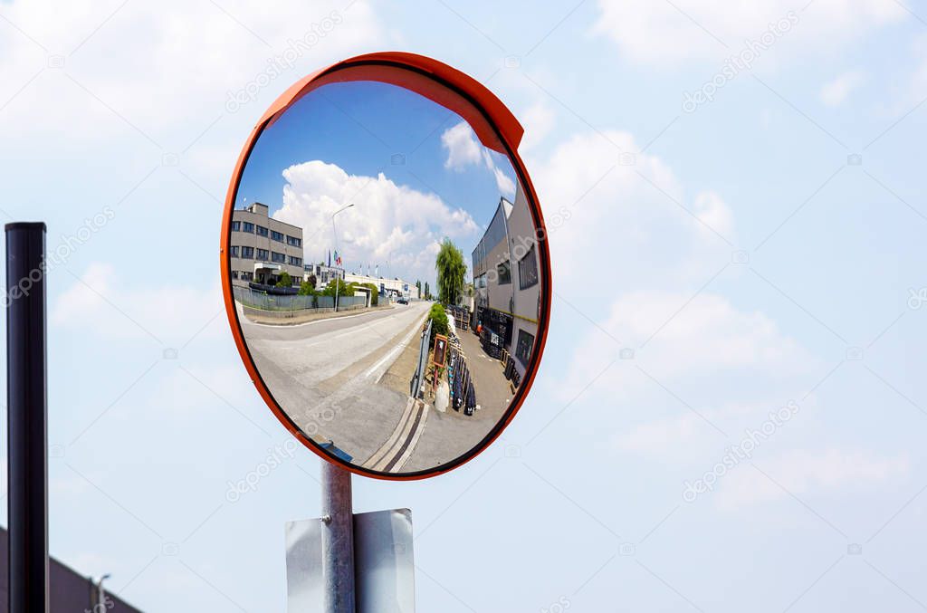 Outdoor convex safety mirror hanging on wall with reflection of an urban roadside view of cars parked along the street by residential apartment buildings.