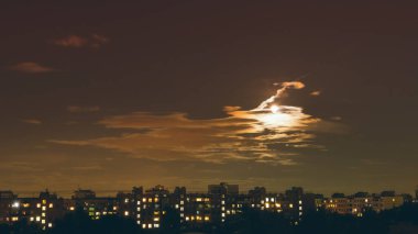 Dolunay, kısmen bulutlarla kaplı, büyük bir şehrin gece konutlarını aydınlatır.