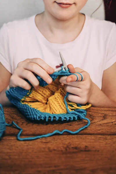 Young girl knitting a circle scarf with yellow and blue coloured yarn sitting at the wooden table. Close up of knitting needles.