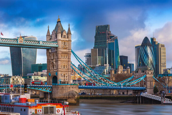London, England - The famous Tower Bridge with iconic red double-decker buses on it and skyscrapers of Bank District at background