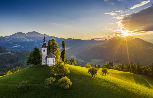 Skofja Loka, Slovenia - Aerial view of the beautiful hilltop Sveti Tomaz (Saint Thomas) church with a warm summer sunset, clear blue sky and Julian Alps at the background