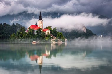Bled, Slovenya - Sisli bir yaz sabahı arka planda, Bled Gölü (Blejsko Jezero) ve bulut kaplı Julian Alps üzerinde yansıması olan küçük bir adada Maria 'nın Varsayımı Kilisesi
