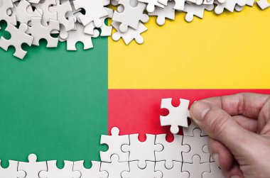 Benin flag  is depicted on a table on which the human hand folds a puzzle of white color.
