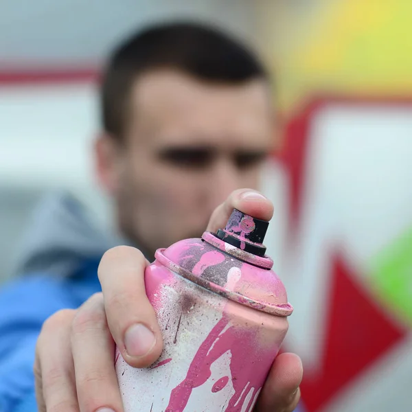 A young graffiti artist in a blue jacket is holding a can of paint in front of him against a background of colored graffiti drawing. Street art and vandalism concept.
