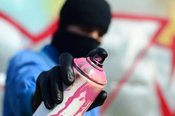 A young graffiti artist in a blue jacket and black mask is holding a can of paint in front of him against a background of colored graffiti drawing. Street art and vandalism concept.