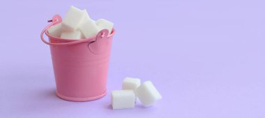 Pembe kova şeker küpleri ile dolu bir minyatür bir Mor pastel arka plan üzerinde yatıyor. En az kavramı.