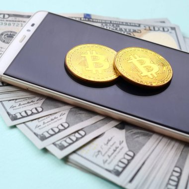 Bir akıllı telefon ve açık mavi arka planı üzerinde yüz dolar faturaları üzerinde Bitcoins yalan.