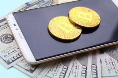 Bir akıllı telefon ve açık mavi arka planı üzerinde yüz dolar faturaları üzerinde Bitcoins yalan.