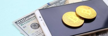 Bir akıllı telefon ve açık mavi arka planı üzerinde yüz dolar faturaları üzerinde Bitcoins yalan.
