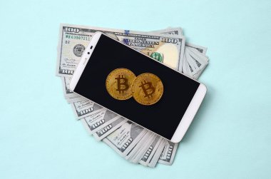 Bir akıllı telefon ve açık mavi arka planı üzerinde yüz dolar faturaları üzerinde Bitcoins yalan.