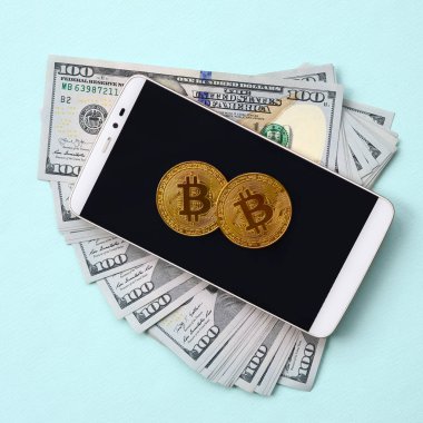 Bir akıllı telefon ve açık mavi arka planı üzerinde yüz dolar faturaları üzerinde Bitcoins yalan.