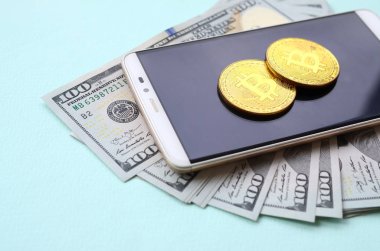 Bir akıllı telefon ve açık mavi arka planı üzerinde yüz dolar faturaları üzerinde Bitcoins yalan.