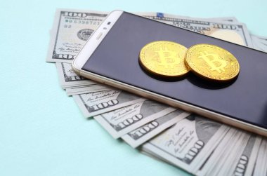 Bir akıllı telefon ve açık mavi arka planı üzerinde yüz dolar faturaları üzerinde Bitcoins yalan.