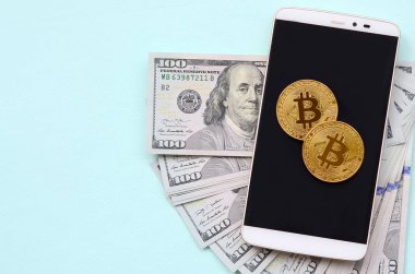 Bir akıllı telefon ve açık mavi arka planı üzerinde yüz dolar faturaları üzerinde Bitcoins yalan.