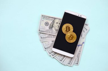 Bir akıllı telefon ve açık mavi arka planı üzerinde yüz dolar faturaları üzerinde Bitcoins yalan.