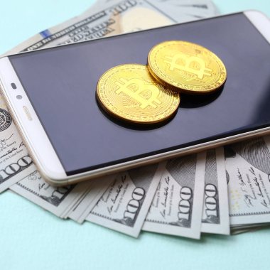 Bir akıllı telefon ve açık mavi arka planı üzerinde yüz dolar faturaları üzerinde Bitcoins yalan.