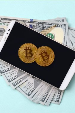 Bir akıllı telefon ve açık mavi arka planı üzerinde yüz dolar faturaları üzerinde Bitcoins yalan.
