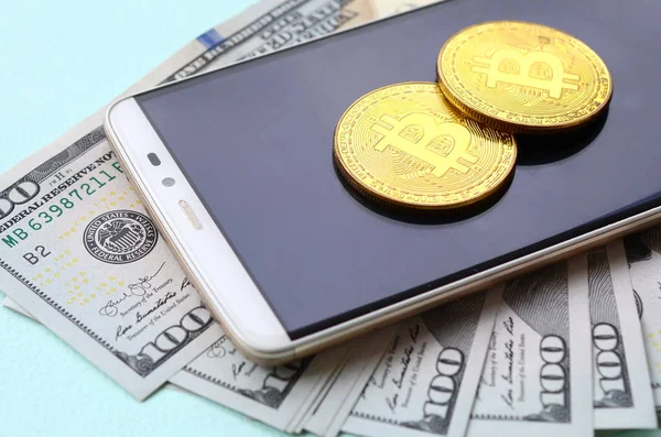 Bir akıllı telefon ve açık mavi arka planı üzerinde yüz dolar faturaları üzerinde Bitcoins yalan.