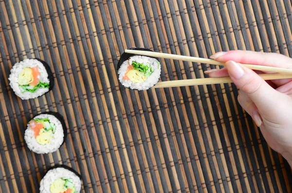 一只拿着筷子的手拿着一个寿司卷放在竹子稻草做的衬垫上 亚洲传统食品 — 图库照片