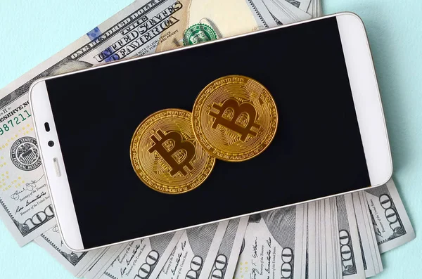 Bir akıllı telefon ve açık mavi arka planı üzerinde yüz dolar faturaları üzerinde Bitcoins yalan.